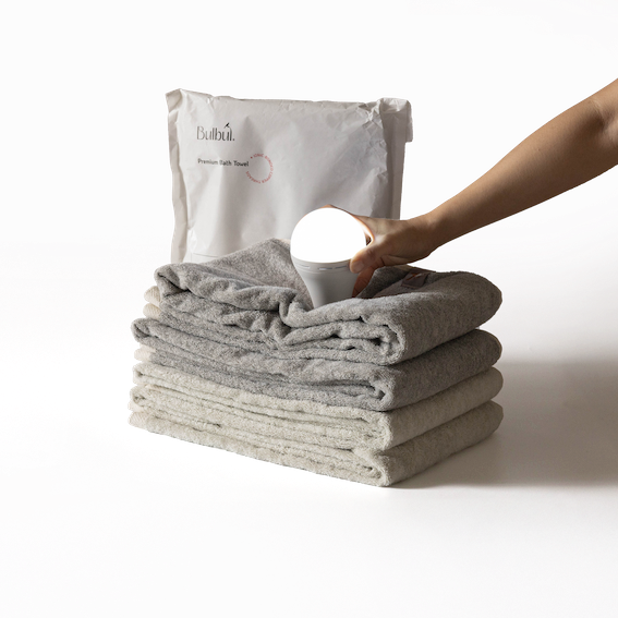 Bulbul Self-sanitizing Premium Copper Bath Towel in grey and beige, stacked neatly with antibacterial packaging and a hand holding a  rechargeable bulb to showcase conductivity because of copper.